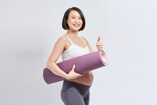 Pregnant girl holding yoga mat standing over white background.