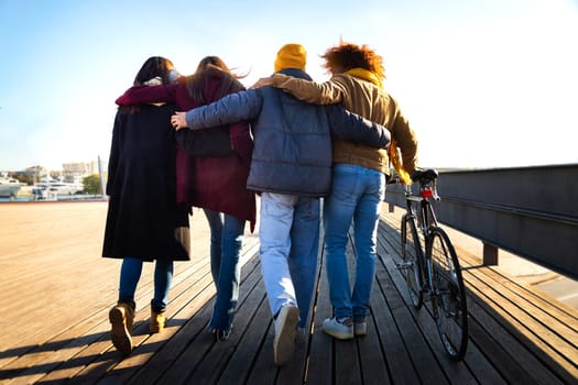 Rear view of group of friends walking together enjoying sunny winter afternoon.Multiracial friends embracing in the city