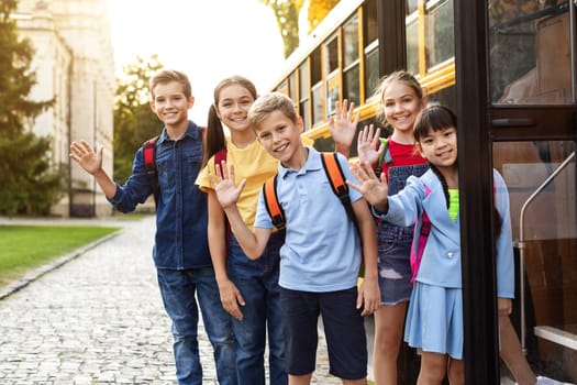 Happy Multiethnic Kids Standing Near School Bus And Waving Hand At Camera, Cheerful Pupils With Backpacks Ready For Trip Home After Lessons, Joyful Children Enjoying School Transportation
