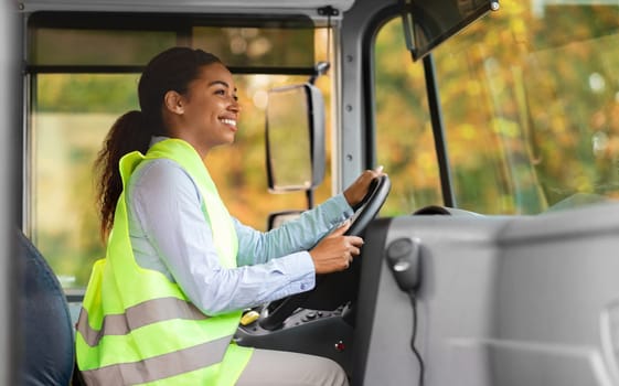 Portrait Of Beautiful Black Driver Woman Driving Public Bus, Happy Young African American Female Wearing Reflective Vest, Holding Steering Wheel And Looking On The Road, Enjoying Her Job, Side View