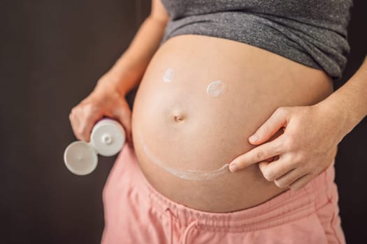 Positive young pregnant woman in comfortable homewear applying belly butter on her big tummy, woman belly