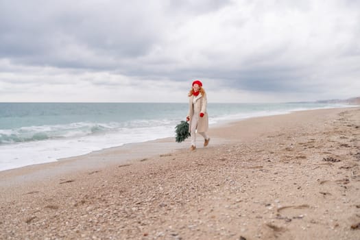 Redhead woman Christmas tree sea. Christmas portrait of a happy redhead woman walking along the beach and holding a Christmas tree in her hands. She is dressed in a light coat and a red beret.
