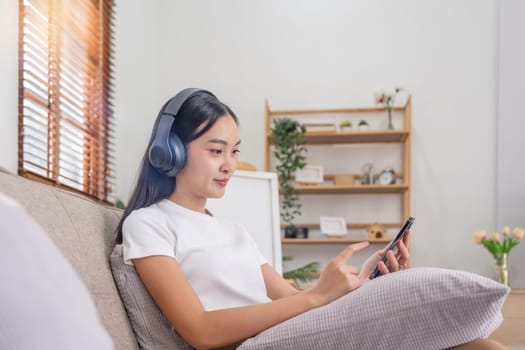 Young Asians woman listen to music on couch in living room at home. Happy Asia female using mobile smartphone, wearing headset and sitting on sofa