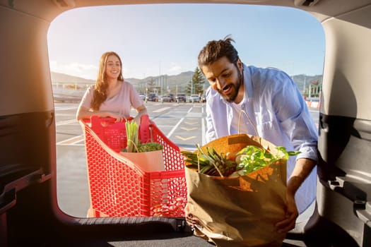 Happy young indian couple packing shopping bags with fresh food into the car trunk, view from the vehicle interior