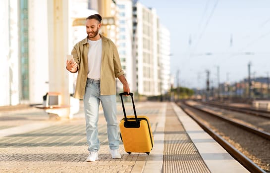 Cheerful young caucasian man with suitcase uses phone, chatting, waiting for transport on train station, full length. Work, travel lifestyle, trip with device app, summer vacation