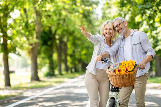 Happy Mature Couple Walking In Park With Retro Bike And Pointing Away, Cheerful Beautiful Senior Spouses Having Fun Together During Outdoor Walk And Showing Copy Space For Advertisement