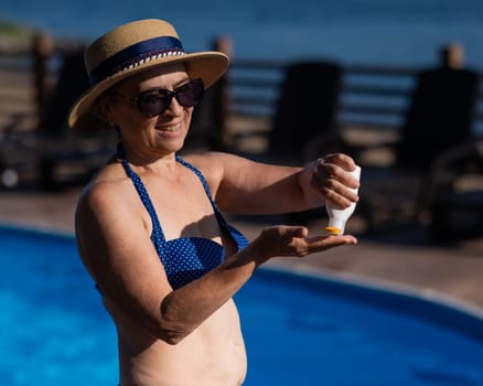 Portrait of an old woman in a straw hat, sunglasses and a swimsuit applying sunscreen to her skin while relaxing by the pool.