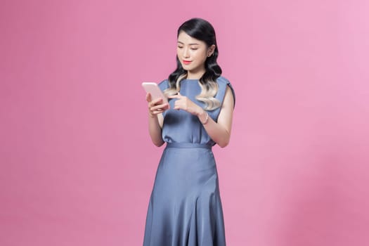 young woman wearing blue silk dress using smartphone against pink background