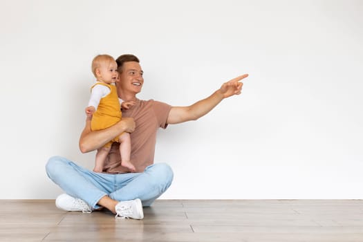 Father With Infant Baby Sitting On Floor Near Wall And Pointing Aside