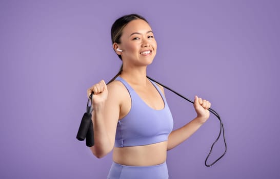 Cardio workout. Smiling asian woman in sportswear holding skipping rope around neck, posing on purple background
