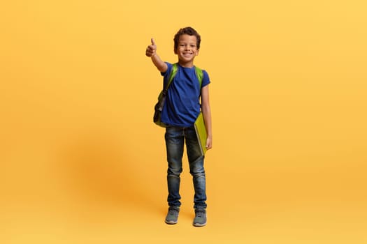 Happy black schoolboy with backpack and exercisebook showing thumb up