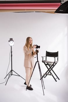 A woman producer, videographer, blogger a blonde with a camera, tripod and light in the photo studio. Wearing a formal nude pantsuit on a white background