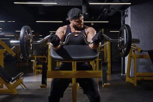 Tattooed athlete in black sport gloves, shorts, vest, cap. Lifting barbell, training his biceps, sitting on preacher curl bench at dark gym. Close up