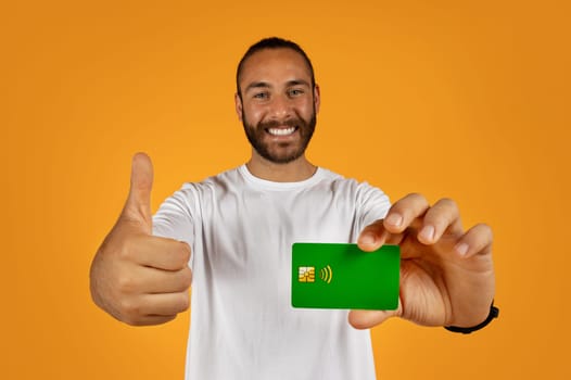 Happy handsome young european guy shows credit card, thumb up, recommends finance savings