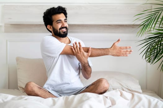 Young Indian Man Stretching Arm Muscles While Sitting In Bed After Awake