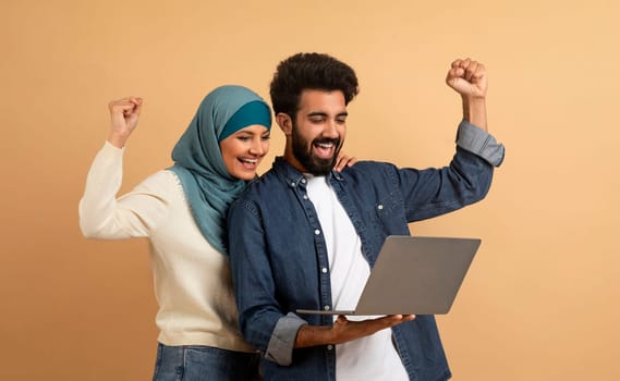 Overjoyed arab couple holding laptop and celebrating success with clenched fists