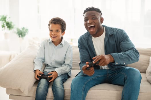 Excited African American Dad And Little Son Playing Game Having Fun Sitting On Sofa At Home. Young Black Father And His Child Play Videogames Together, Spending Good Time On Weekend
