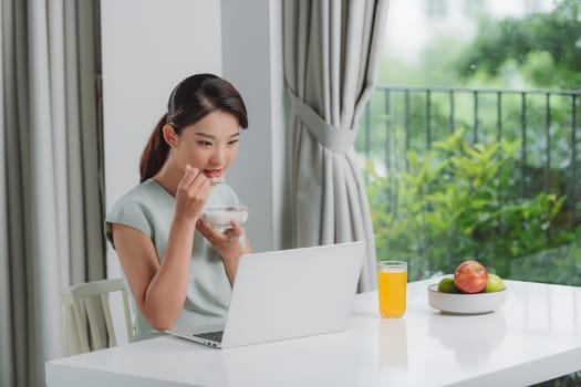 Satisfied woman eating breakfast using a laptop on a desk at home