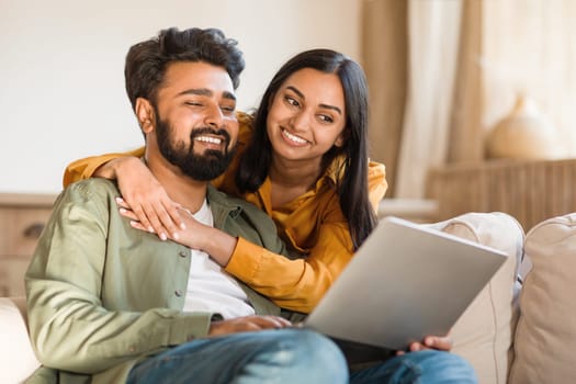 Happy young indian couple using laptop at home together, shopping online or booking vacation, resting on couch