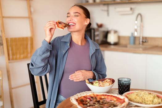 Young hungry pregnant woman biting slice of pizza, having desire for junk food, sitting at table full of dishes