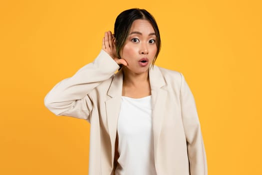 Shocked curious young asian woman in suit, put hand to ear, listening to gossip