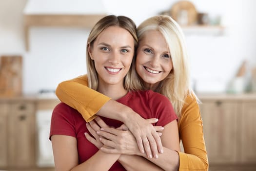 Loving attractive blonde senior woman mother hugging her young adult daughter. Happy cheerful elderly and millennial ladies embracing at home, posing together for family photo at cozy kitchen
