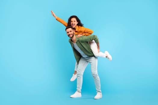 Handsome man giving piggyback ride to his wife against blue studio background, full length portrait, free space