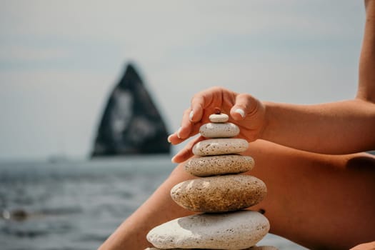 Woman building a stone pyramid on the seashore on a sunny day. Blue sea background. Travel destination, relaxation and meditation concept.