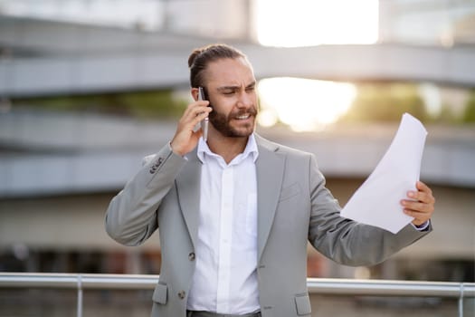 Business Problems. Angry Young Businessman Holding Papers And Talking On Cellphone Outdoors, Furious Male Entrepreneur Standing Outside Near Modern Office Building, Managing Issues By Phone
