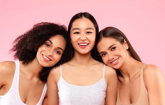 Happy three diverse female friends models posing and smiling to camera, standing on pink background, studio shot