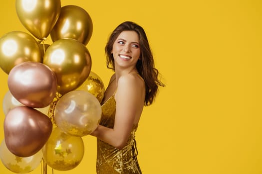 Party time. Happy european woman in elegant dress holding bunch of birthday balloons over yellow studio background, looking back at copy space, having holiday celebration