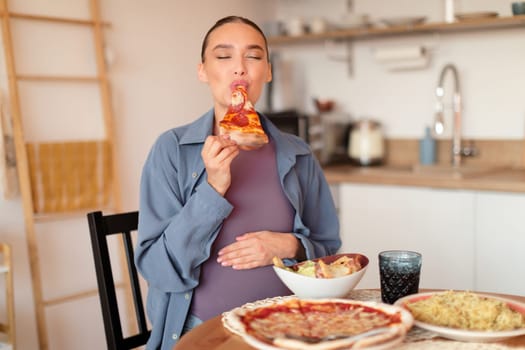 Pregnancy and cheat meal. Young expectant woman having desire for junk food, biting slice of pizza, sitting in kitchen
