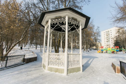 Beautiful white gazebo in a winter park against the backdrop of snowy trees. Park in city.