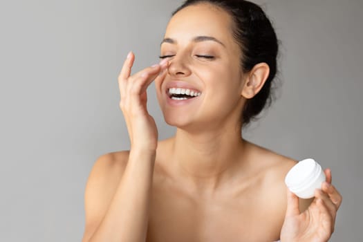 Happy woman applying moisturizer cream on nose on gray background
