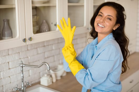 Portrait of joyful woman putting on rubber gloves, ready to tidy house or washing dishes standing in kitchen, free space