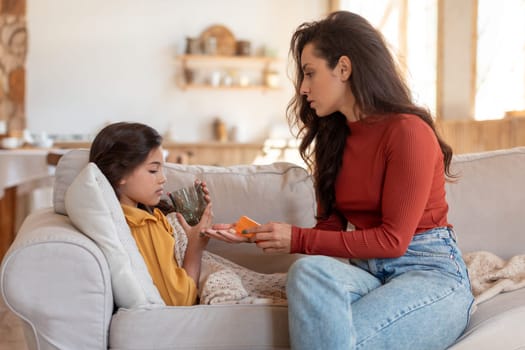 Flu Treatment. Young mother giving a glass of water and pill to her sick daughter on couch at home. Kid girl taking medication during influenza illness. Child health problems concept