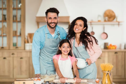Portrait of happy european family of three posing while cooking pastry in kitchen together, smiling at camera