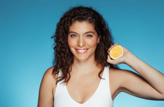 Glad millennial european curly lady show orange fruit, isolated on blue studio background. Lifestyle, vitamin C