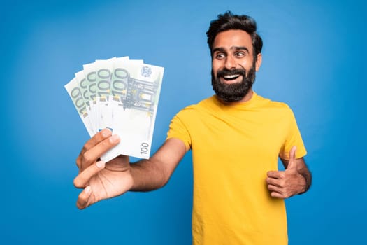 Excited rich indian middle aged man holding euro money and gesturing thumb up, celebrating big financial luck
