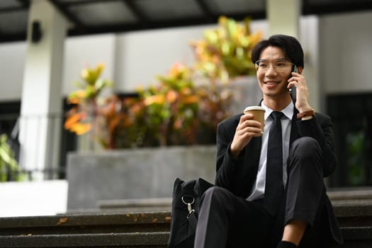 Smiling male worker drinking coffee and talking on mobile phone outside office building