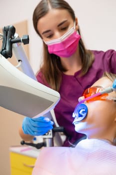 A dentist uses a saliva ejector while conducting a professional teeth whitening procedure for a young female patient, vertical photo