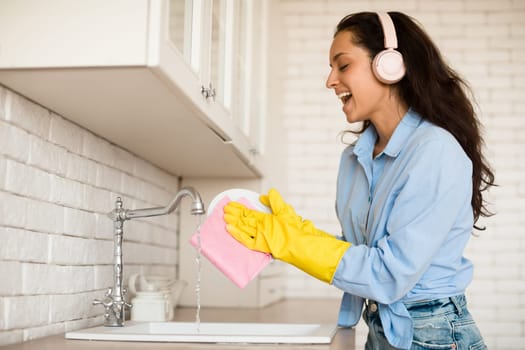 Joyful woman wearing wireless headphones and singing while washing dishes in kitchen, enjoying making domestic chores