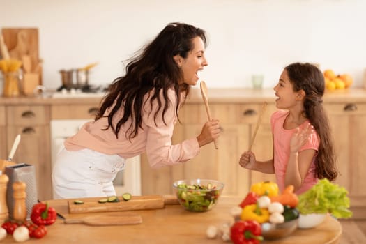 Cooking Fun. Positive Mother And Kid Daughter Singing While Preparing Dinner, Holding Spoons Like Microphones In Modern Kitchen Indoor. Family Fooling While Making Salad Together