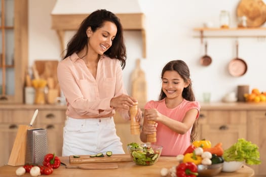 Family Nutrition. Happy Mother And Preteen Daughter Cooking And Seasoning Salad, Adding Salt And Pepper Spices To Food, Preparing Healthy Vegetable Meal For Dinner At Modern Kitchen Interior