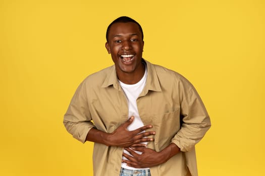 Laughing millennial african american guy in casual clothes presses hands to stomach