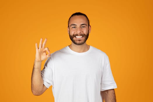 Cheerful adult european man with beard in white t-shirt making ok sign with hand