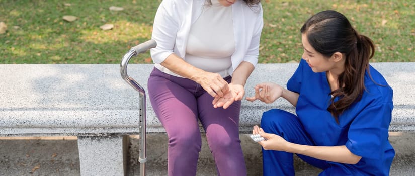 Caregiver or nurse giving and holding medicine pill to senior woman on park chair
