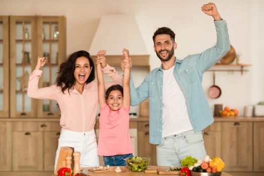Joyful family holding hands and shaking fists, gesturing yes while preparing dinner together in kitchen