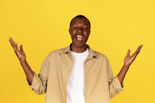 Glad millennial african american man in casual raises hands, rejoices to success, celebrates win