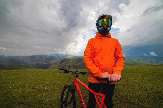 cyclist in an orange bright jacket and a protective helmet stands and holds a bicycle in the mountains, outside the city, off the road in summer in cloudy weather
